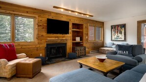Un salon chaleureux, aménagé dans le style d’un chalet, avec une cheminée, des murs en bois et une grande fenêtre.