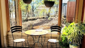 Une petite terrasse avec deux chaises et une table, un pot de fleurs et une vue sur un jardin, visible à travers une fenêtre.