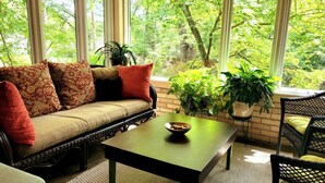 Un coin salon confortable, avec un canapé, une table basse et des fauteuils, entouré de grandes fenêtres donnant sur un paysage verdoyant.