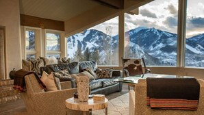 Ein gemütliches Wohnzimmer mit Ledersofa, einem gemusterten Sessel und einem Couchtisch. Große Fenster bieten Blick auf schneebedeckte Berge.