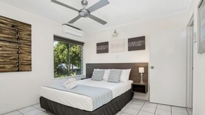 A bedroom with a bed, a ceiling fan, a window with a view of greenery, and wall-mounted artwork. - Seascape Holidays at The Queenslander Two Bed Standard (Port Douglas)