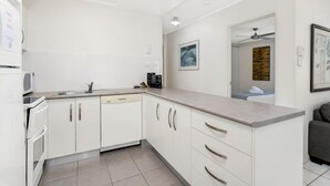 A modern kitchen with white cabinets, a countertop, and a microwave. There is a ceiling fan in the adjacent room. - Seascape Holidays at The Queenslander Two Bed Standard (Port Douglas)