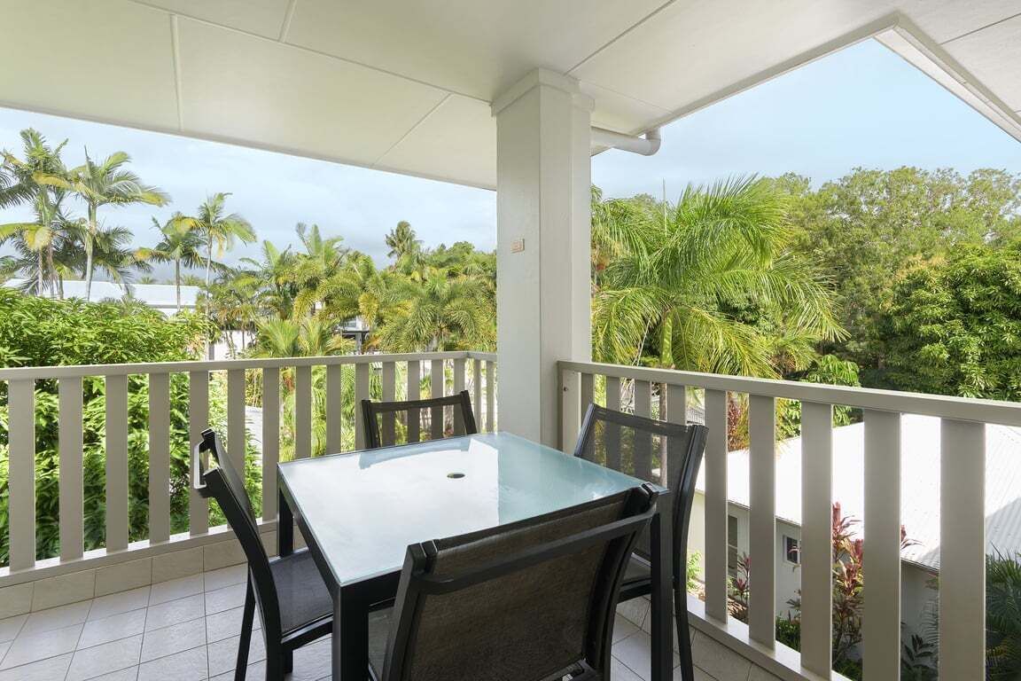 A covered balcony with a glass table and chairs, surrounded by white railings, overlooking a lush green landscape with palm trees.