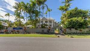 A hotel with a white facade, palm trees, and a fenced area with flowers. - Seascape Holidays at The Queenslander Two Bed Standard (Port Douglas)