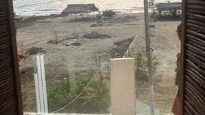 Vista a la playa desde una ventana, con el mar, cabañas de playa y algo de vegetación.