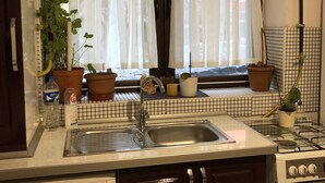 A kitchen with a built-in washing machine, a sink, and a stove. There are potted plants on the windowsill and a window with sheer curtains. - Mavi Ladin Konukevi (Kars)