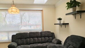 En stue med en mørkegrå hjørnesofa, en matchende lænestol, en hængende lampe, en potteplante på en hylde og et vindue med persienner.