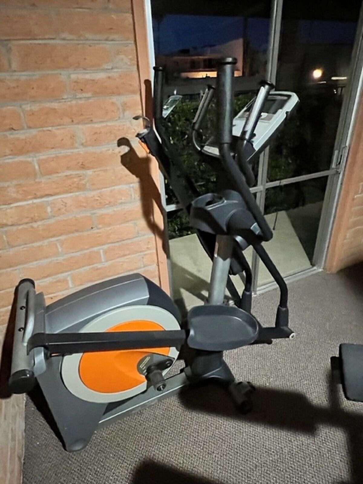 A stationary exercise bike with a grey and orange design, positioned indoors with a brick wall and a glass door leading to an outdoor area.