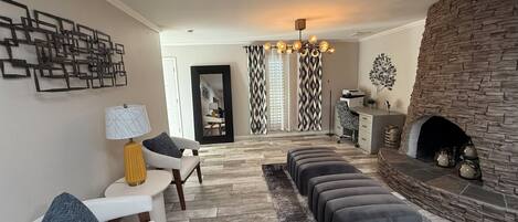 A living room with a fireplace, a dining area, and a seating area with ottomans and a lamp.