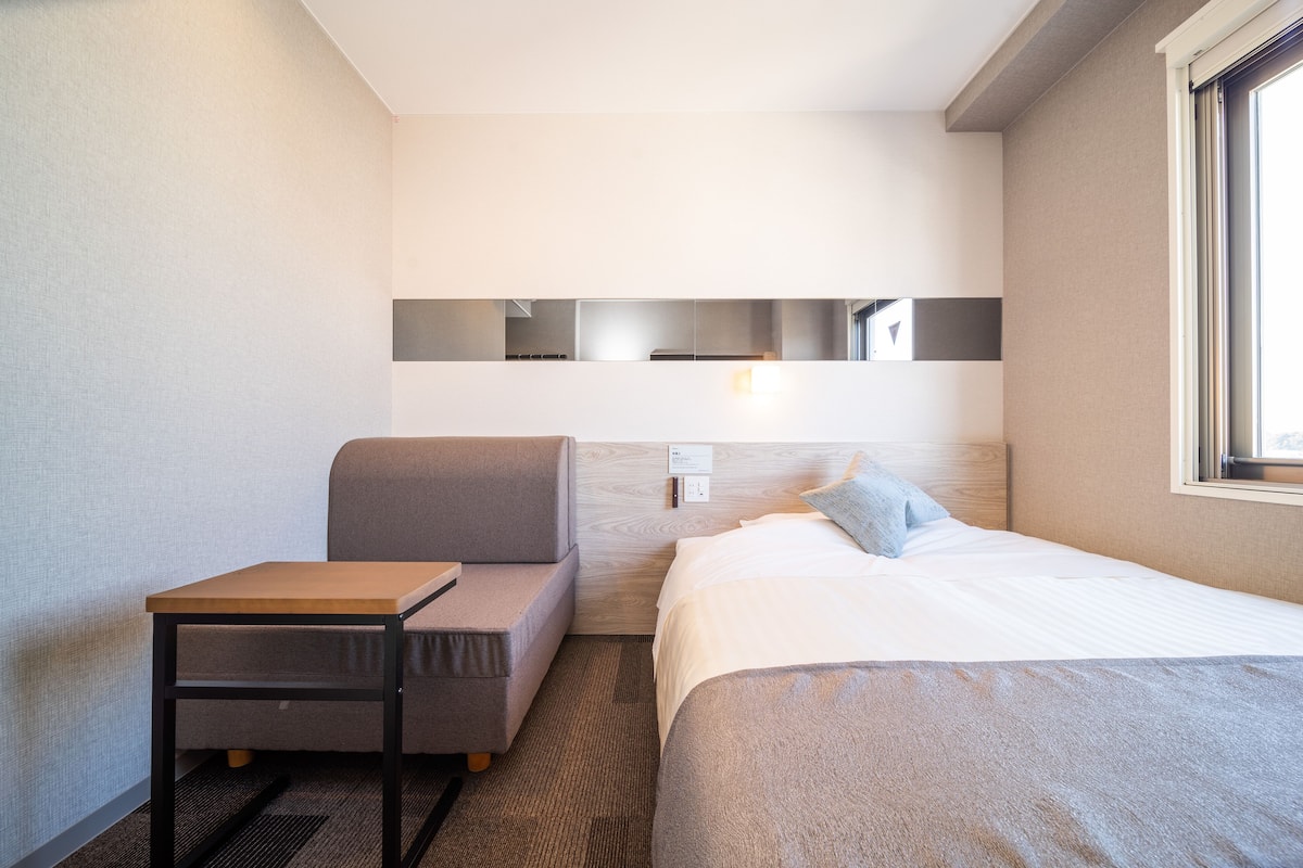 Ein modernes Hotelzimmer mit Bett, kleinem Tisch, Sofa und Wand-Fernseher.