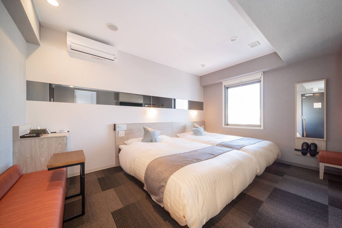 Ein modernes Hotelzimmer mit zwei Betten, einem Schreibtisch, einer Bank und einem großen Fenster.