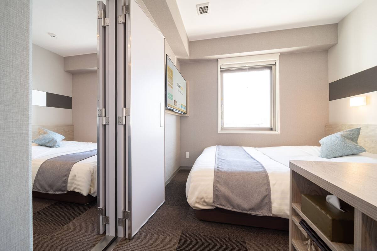 Ein Hotelzimmer mit zwei Betten, einem Fenster, einem Schreibtisch und einem an der Wand befestigten Fernseher.