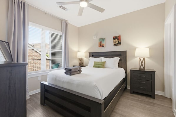 A bedroom with a bed, two bedside tables with lamps, a ceiling fan, and artwork on the wall.