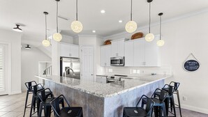 Una cucina moderna con mobili bianchi, elettrodomestici in acciaio inossidabile, un'isola in marmo con sgabelli da bar e illuminazione a sospensione.