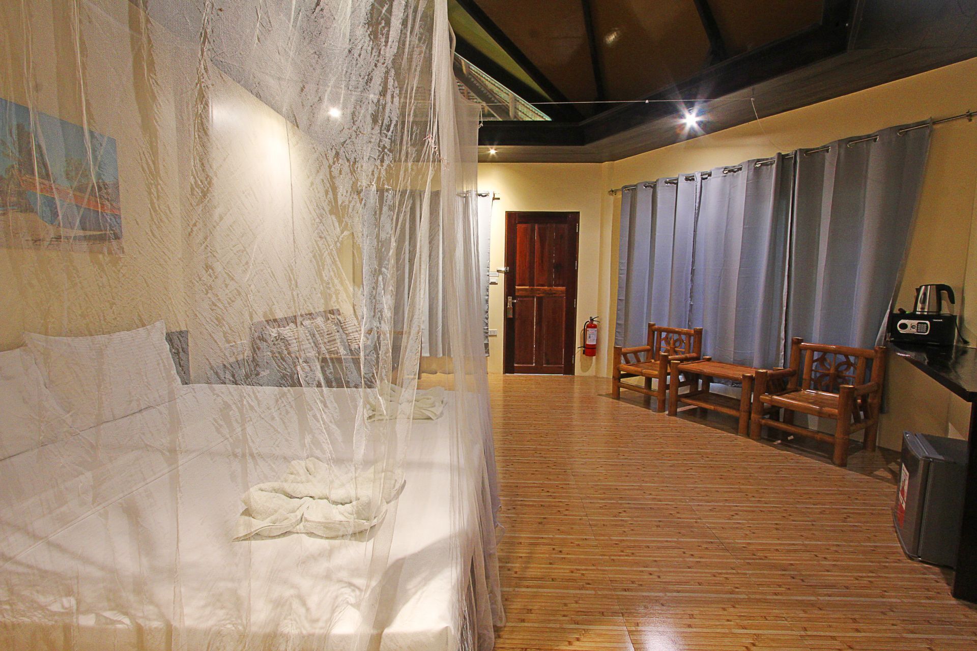 A hotel room with a bed, wooden floor, curtains, a table with chairs, a coffee maker, and a fire extinguisher.