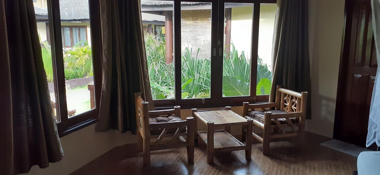 A room with a wooden table and chairs, large windows with curtains, and a view of greenery outside.
