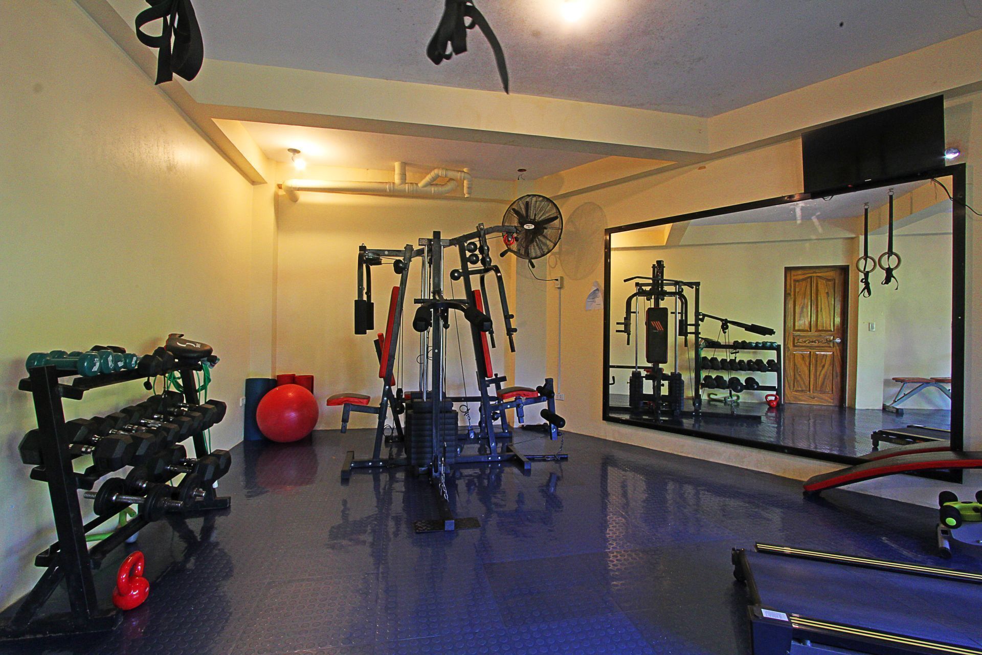 Uma academia com diversos equipamentos de exercícios, um espelho grande e uma porta com moldura de madeira.