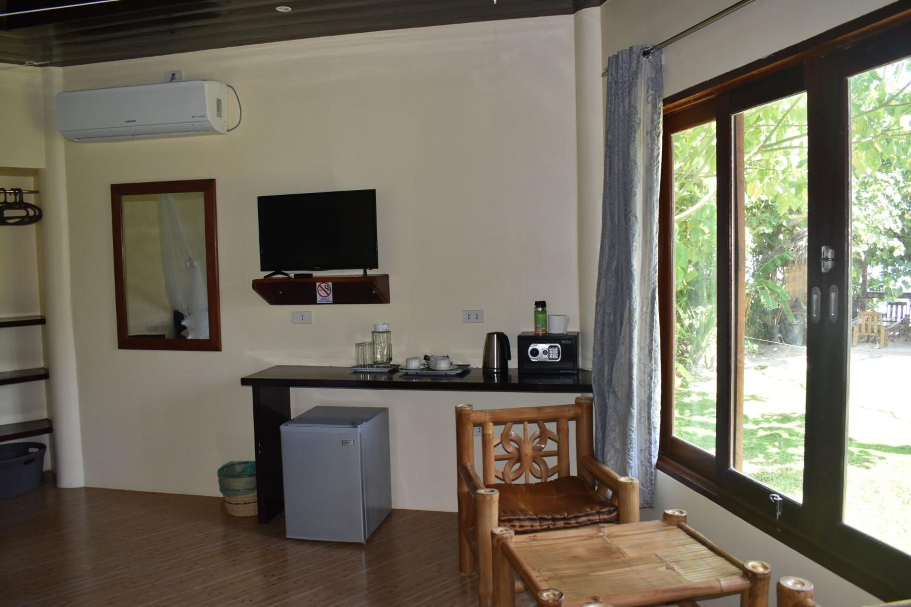 A compact room with a kitchenette, a television, a small refrigerator, a wooden chair, and a large window with a view of greenery.