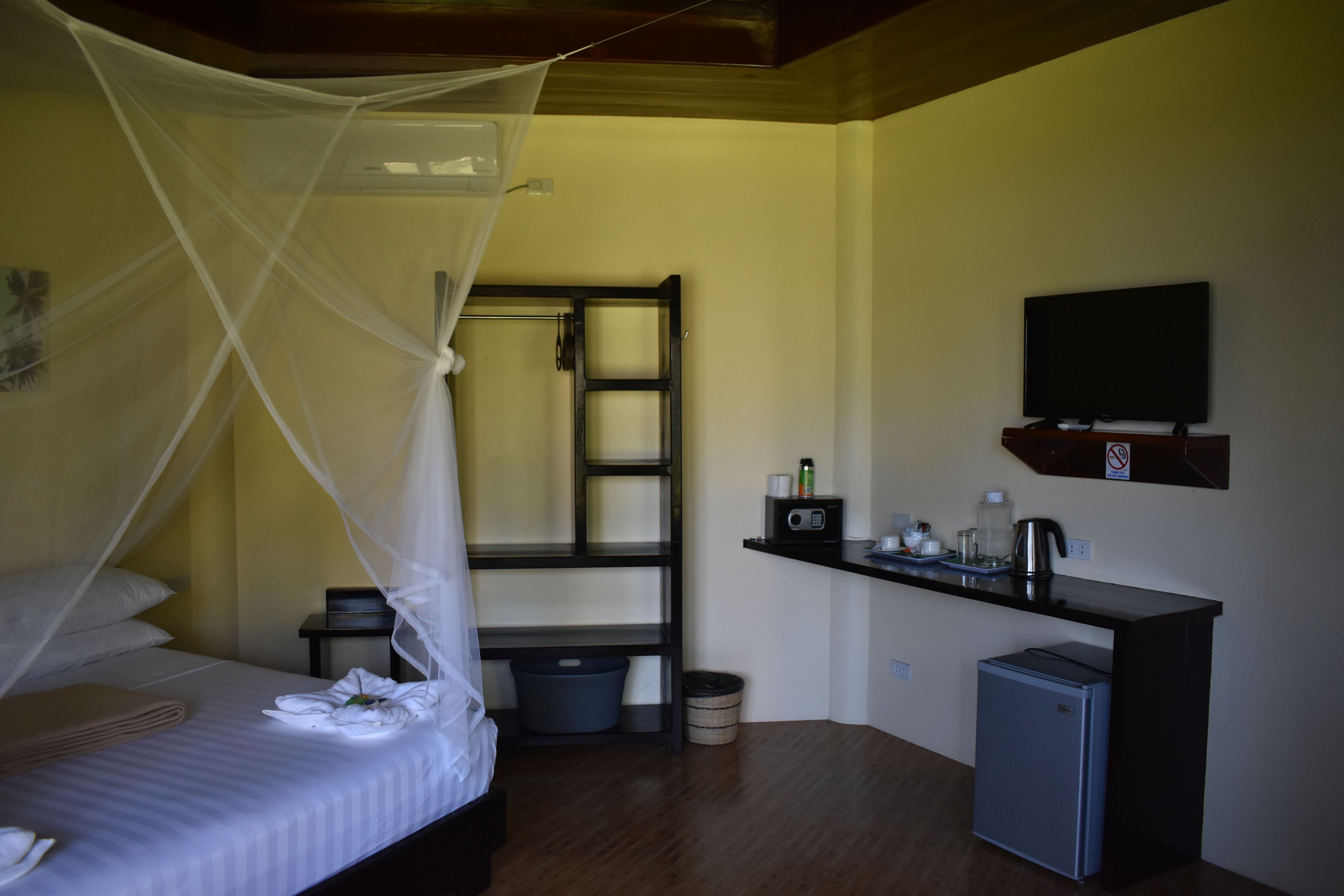 A bedroom with a bed, mosquito net, bedside table, and a small kitchenette area with a TV, radio, and refrigerator.