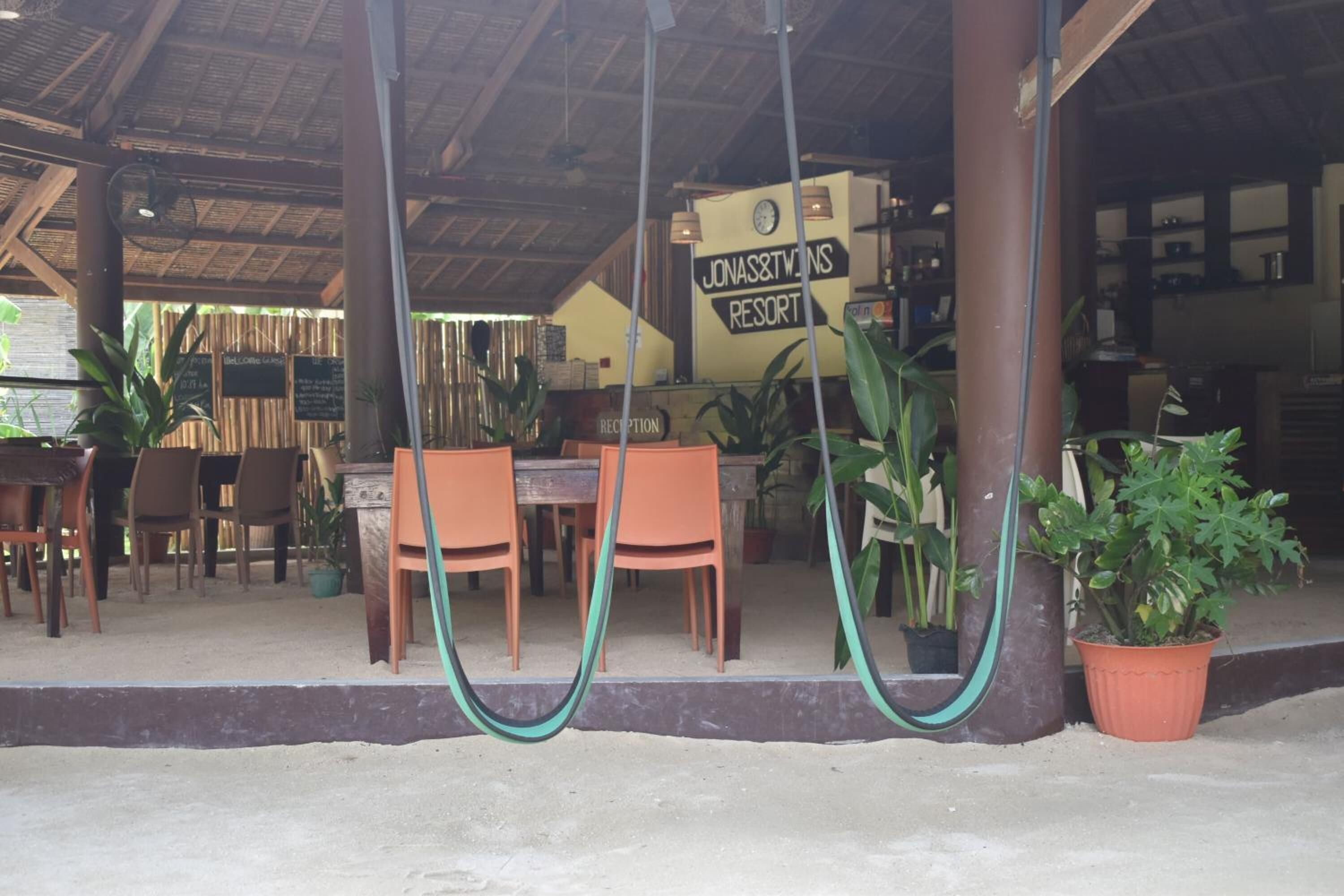 Un área de recepción con un cartel que anuncia Jonas & Twins Resort, mesas y sillas de madera, y plantas en macetas.