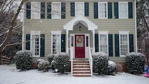 Une maison à deux étages avec une porte rouge et une couronne décorative, entourée d’arbustes couverts de neige et d’une pelouse enneigée.
