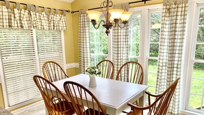 Une salle à manger avec une table blanche et des chaises en bois, de grandes fenêtres équipées de stores et un lustre.