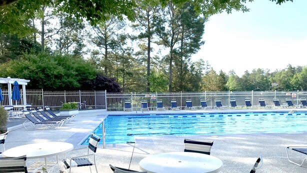 Une piscine entourée d’une clôture et d’arbres, avec des chaises longues et des tables à proximité.