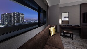 Habitación de hotel moderna con un amplio ventanal que ofrece vistas a la ciudad, un sofá cómodo, un escritorio con silla y un televisor de pantalla plana.