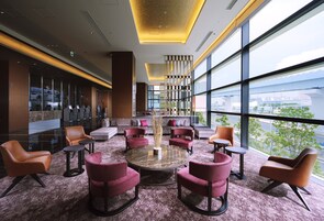 A modern lounge area with a central round table surrounded by chairs, large windows offering a city view, and a decorative pillar in the center.