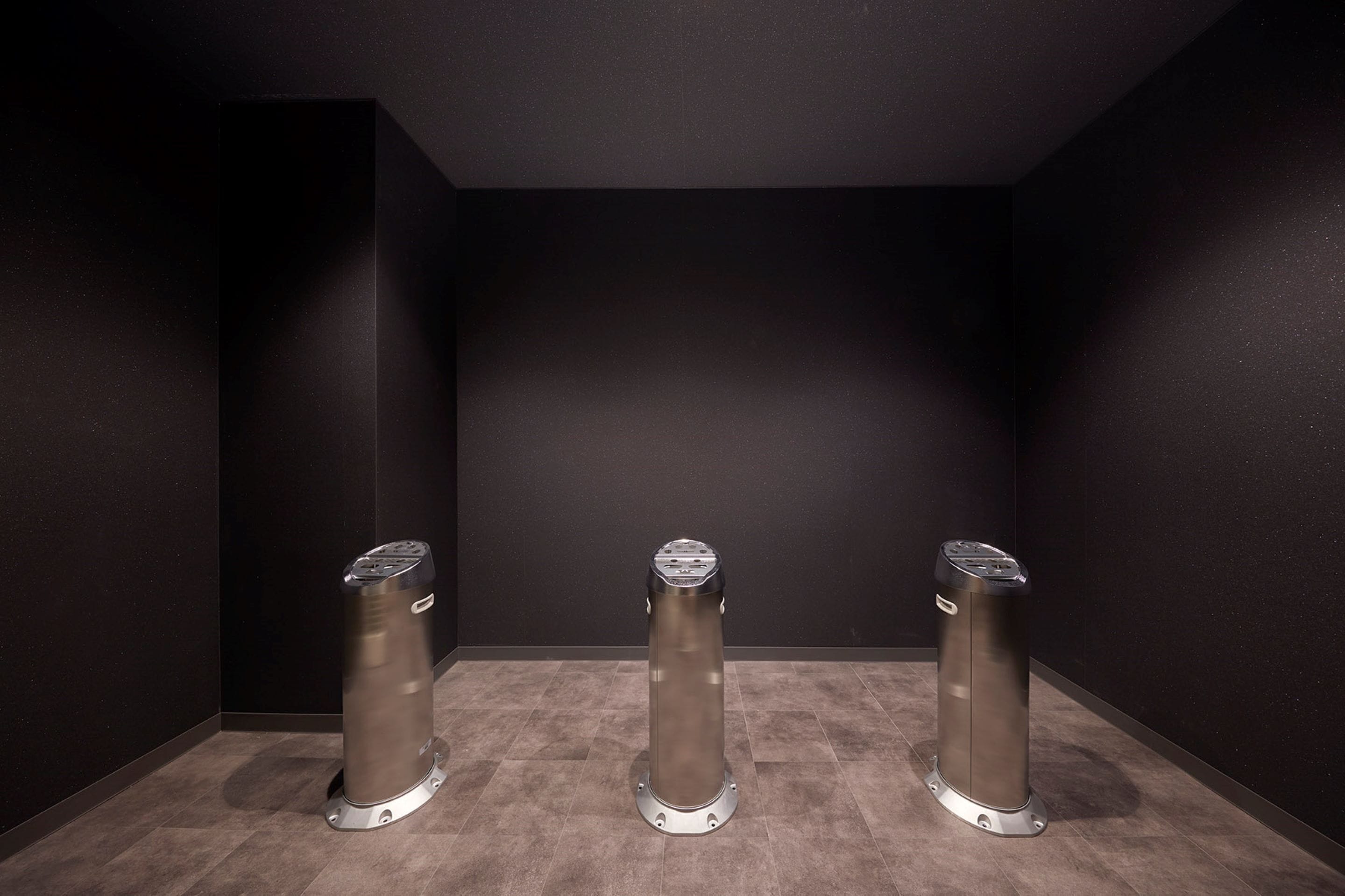 Trois poubelles cylindriques sont placées dans un coin d’une pièce aux murs sombres et au sol carrelé.
