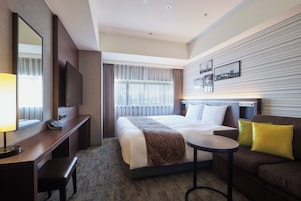 A hotel room with a bed, a desk, a chair, a small table, a sofa, and a TV.