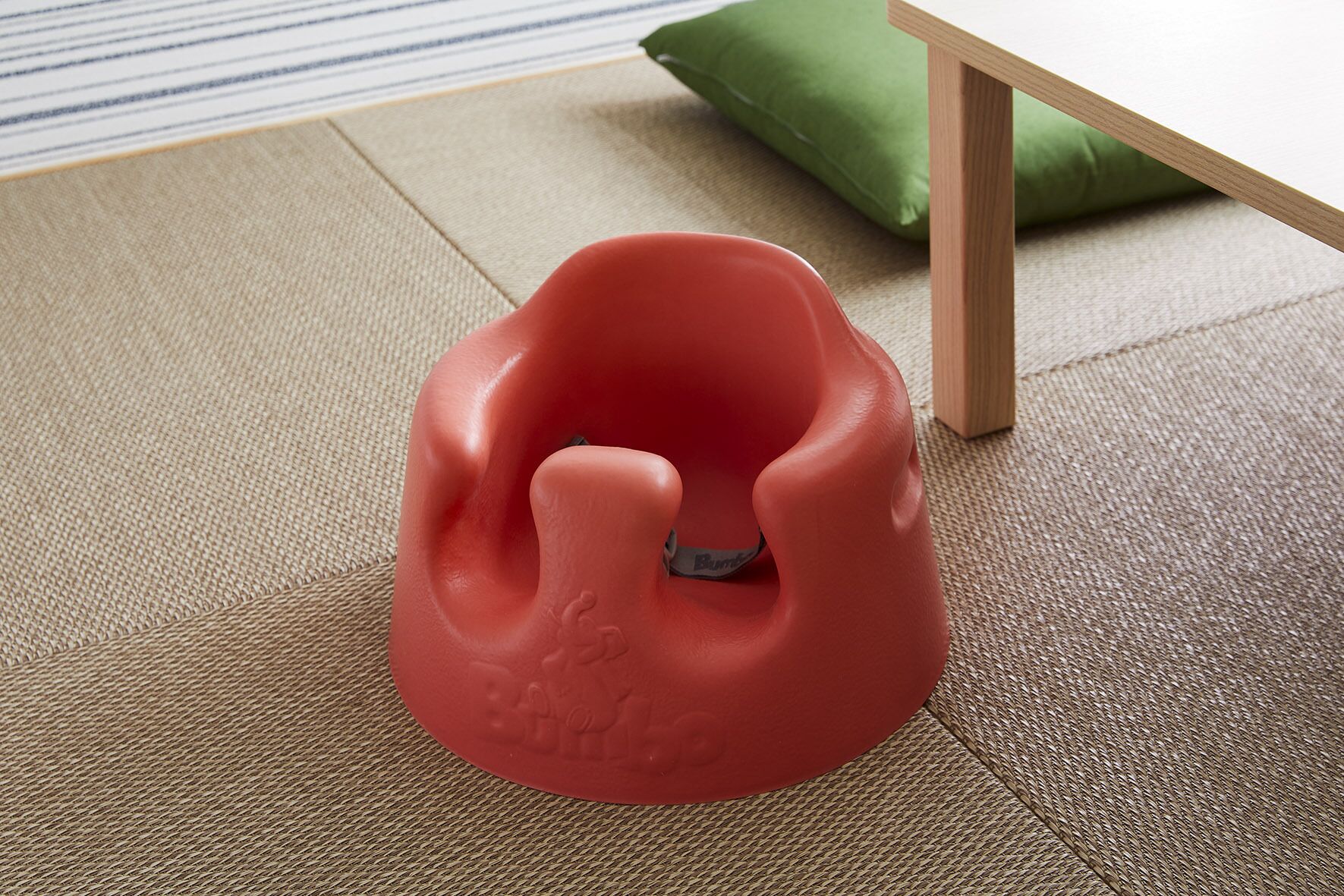 Un objeto rojo y texturizado sobre un suelo tejido, con un cojín verde y una mesa de madera al fondo.