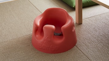 Un objeto rojo y texturizado sobre un suelo tejido, con un cojín verde y una mesa de madera al fondo.