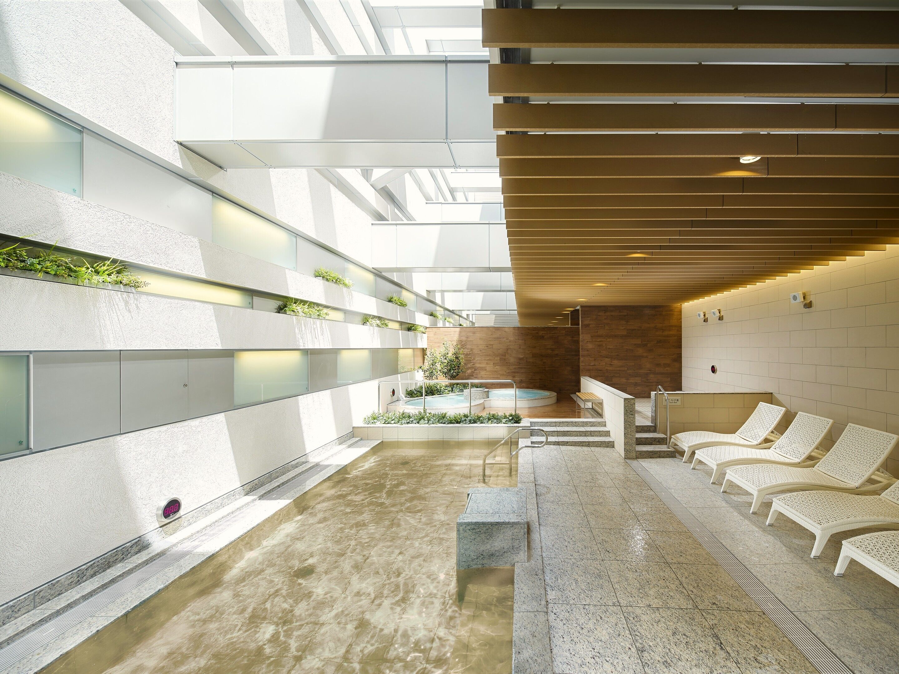 Un espace piscine intérieur moderne, doté de murs en verre transparent, d’un élément décoratif central avec de l’eau et de chaises longues disposées le long du pourtour.
