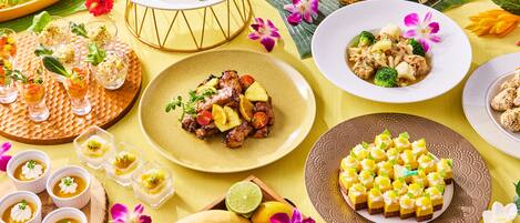 A variety of dishes and desserts on a table with a yellow tablecloth, accompanied by tropical flowers and fruits.