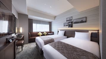 A hotel room with two beds, a desk, a chair, a TV, and a sofa with cushions.