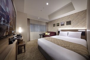 A hotel room with a large bed, a desk with a computer, a TV, and a view of the city through the window.