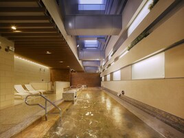 A modern indoor pool area with clear glass ceilings, sunlit walls, and a central water feature.