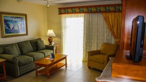 A living room with a sofa, armchair, coffee table, lamp, television, and a painting on the wall.