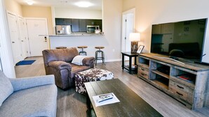 A living room with a sofa, ottoman, coffee table, TV stand, and kitchen area in the background.