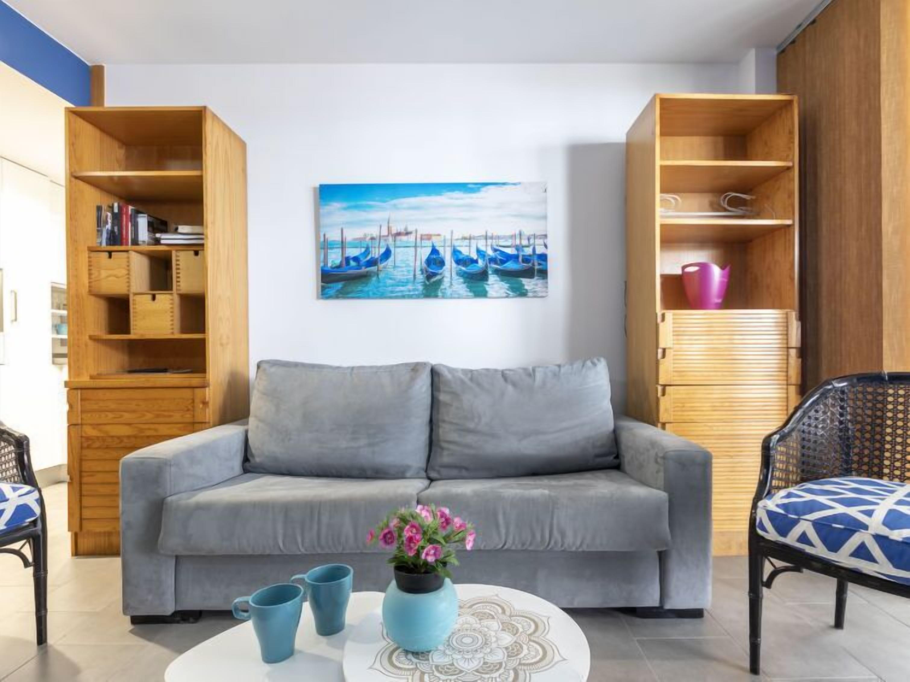 A modern living room with a grey sofa, a coffee table with a vase of flowers, and a painting of boats on the wall.