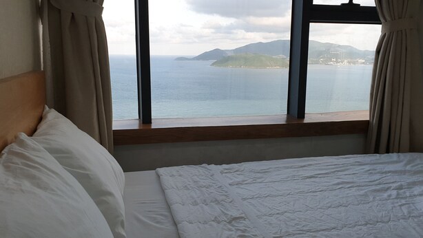 A hotel room with a bed, a window with curtains, and a view of the sea and distant islands. - Nina Apartments (Nha Trang)