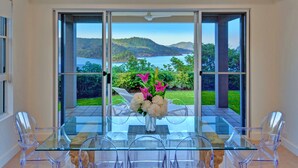 A glass dining table with clear chairs, a vase of flowers, and a scenic view of a lake and hills through large sliding glass doors. - La Bella Waters 1 Wide Ocean Views (Whitsundays)