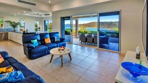 A spacious living room with a blue sofa, a coffee table, and a view of a lake through large sliding doors. - La Bella Waters 1 Wide Ocean Views (Whitsundays)