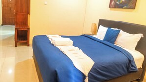 A neatly made bed with blue and white bedding, a wooden nightstand with a lamp, and a painting on the wall. - Kuta Paradise 777 (Legian)