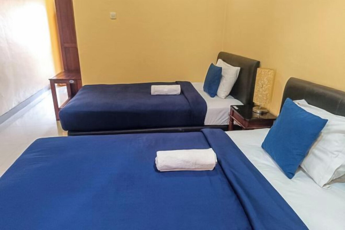 A hotel room with two beds, each with blue bedding and white pillows. There is a small wooden nightstand with a lamp between the beds.