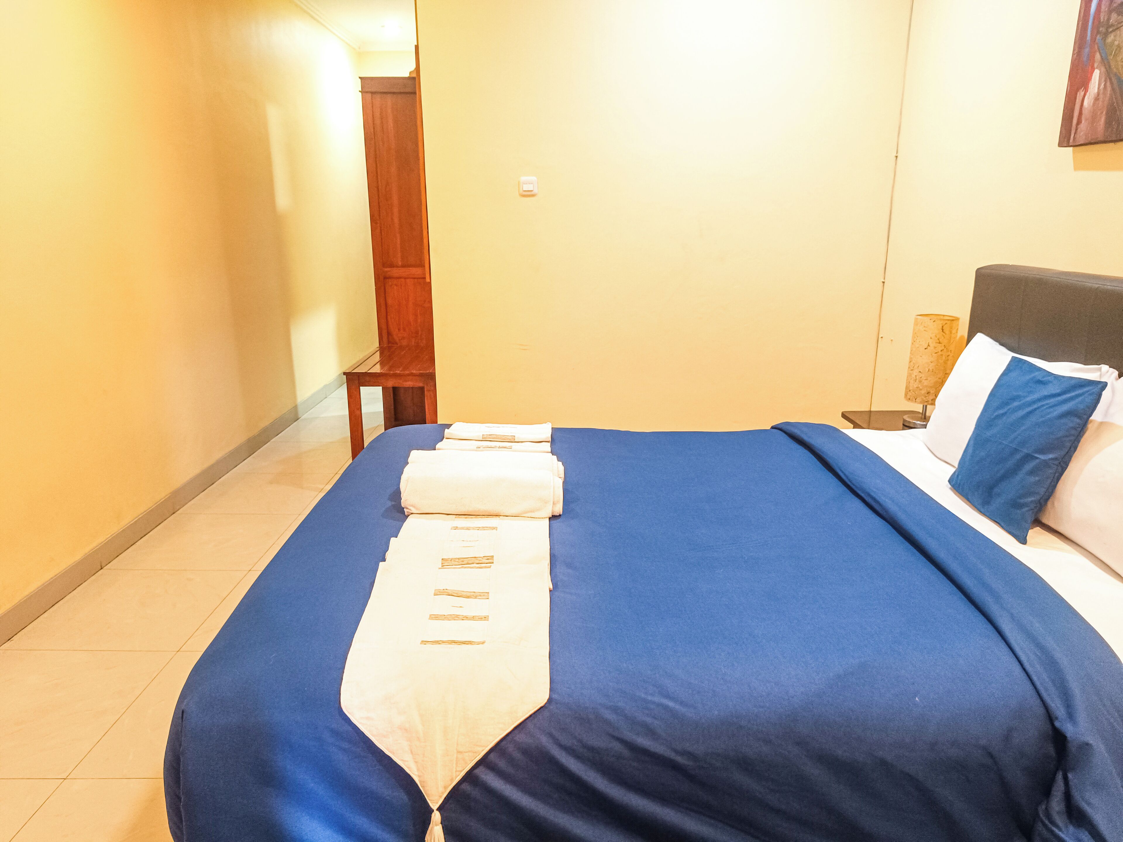 A hotel room with a neatly made bed, a wooden nightstand, a lamp, and a painting on the wall.