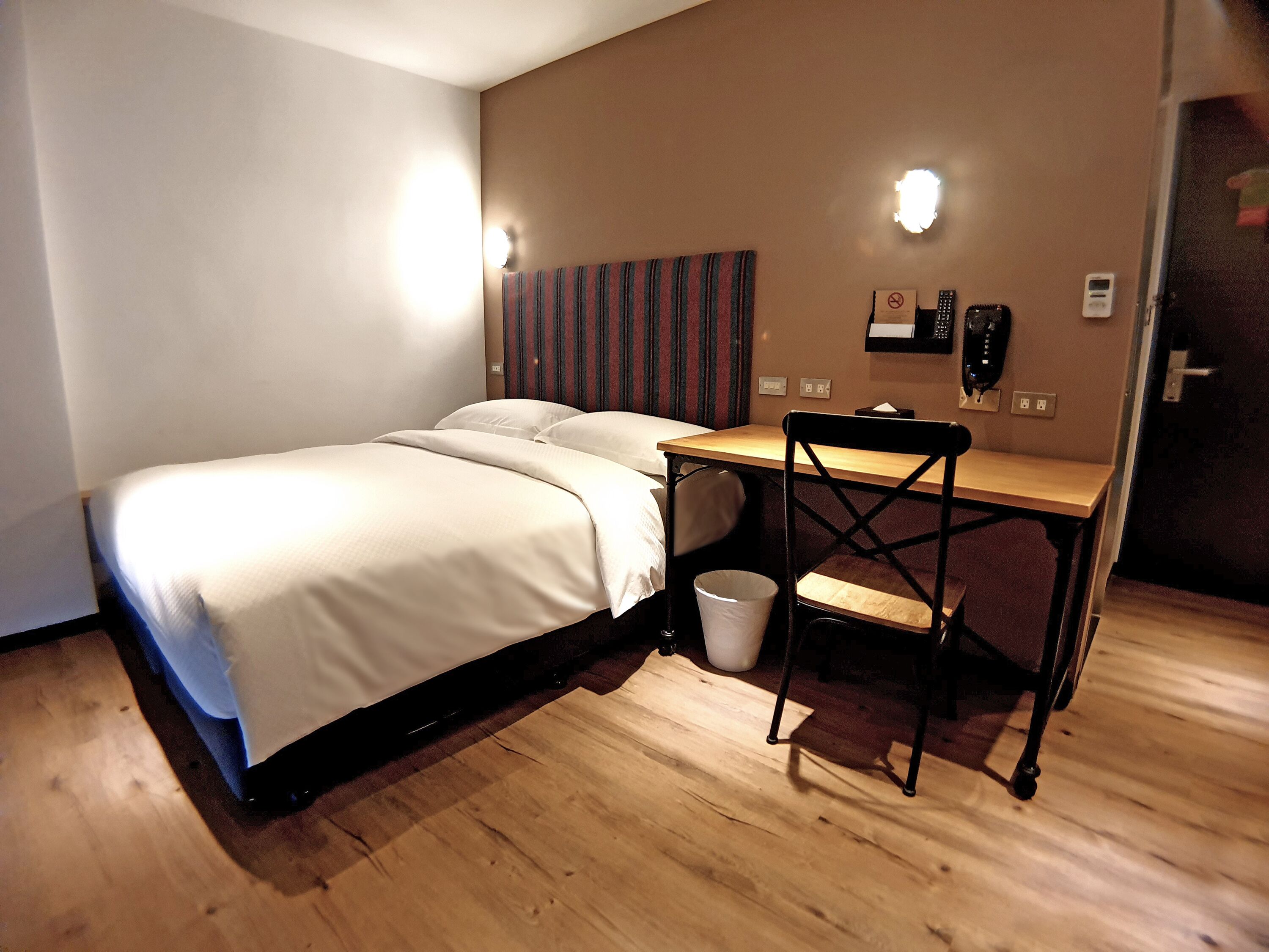 Une chambre d’hôtel avec un lit, un bureau et une chaise. Il y a deux lampes fixées au mur et un téléphone accroché au mur.