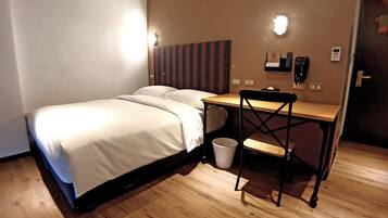 Une chambre d’hôtel avec un lit, un bureau et une chaise. Il y a deux lampes fixées au mur et un téléphone accroché au mur.