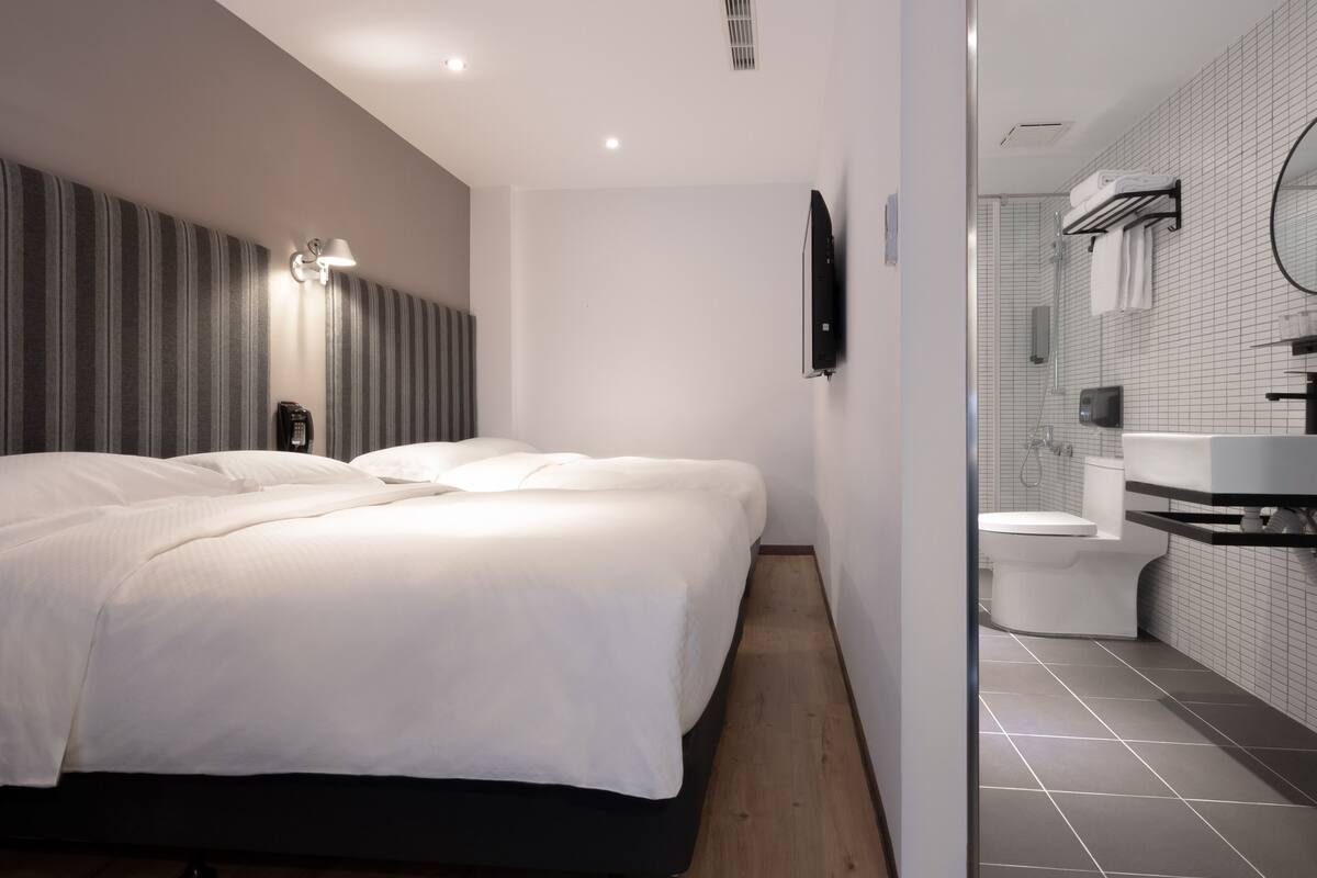 Quarto de hotel moderno com uma cama grande, mesa de cabeceira, televisão de tela plana e banheiro com vaso sanitário, pia e espelho.